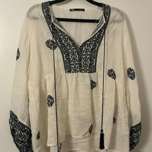Vit och svart blus från Zara - Vit blus från Zara med svarta broderade detaljer och mönster. Långärmad med lös passform, v-ringning och knytband med tofsar. Tillverkad i ett lätt bomullstyg, perfekt för en chill och bohemisk vibe.