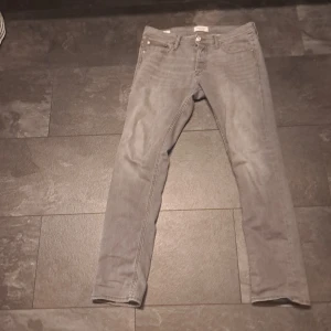 Grå jeans från Jack & Jones - Snygga grå jeans från Jack & Jones. Klassisk femficksmodell med knappgylf och diskreta sömmar. Jeansen har en stilren look och passar perfekt till sneakers eller boots. Tillverkade i mjukt denimtyg för skön känsla hela dagen.
