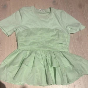 Mintgrön volangblus från Day + Moon - Superfin mintgrön blus från Day + Moon med korta ärmar och rund hals. Toppen har markerad midja och volangig peplum nedtill som ger en söt och trendig siluett. Perfekt för dig som gillar pastell och vill ha något unikt i garderoben. Aldrig använd!