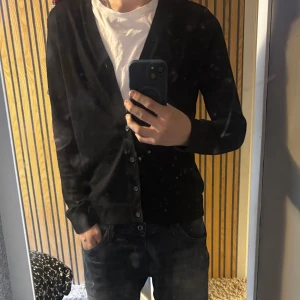 Svart merinoullskofta från H&M - Svart kofta från H&M i mjuk merinoullsblandning. Klassisk modell med v-ringning, knappar framtill och långa ärmar. Perfekt att slänga över en t-shirt för en clean och stilren look.