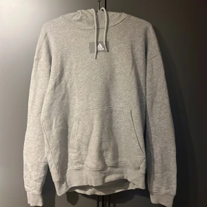 Grå hoodie från Adidas - Säljer en stilren grå hoodie från Adidas med klassisk huva och snörning. Tröjan har en stor magficka och ribbade muddar vid ärmslut och nederkant. Perfekt för en avslappnad look och enkel att matcha med det mesta.