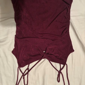 Vinröd linnetopp med snörning - Säljer en vinröd linnetopp med smala axelband och snygg snörning framtill. Toppen har en tight passform och är tillverkad i ett mjukt, stretchigt material som sitter skönt mot kroppen. Perfekt för dig som vill ha en trendig och stilren look. Själv köpt linnet second hand men aldrig använt det. Fler bilder kan skickas om önskas och pris går att diskutera.