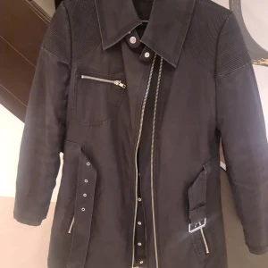 Svart lång kappa med bälte och dragkedjor - Snygg svart lång kappa med coola detaljer som bälte i midjan, flera dragkedjor och quiltat foder. Jackan har stora fickor, markerad krage och förstärkta axelpartier. Perfekt för dig som gillar en stilren men edgy look.