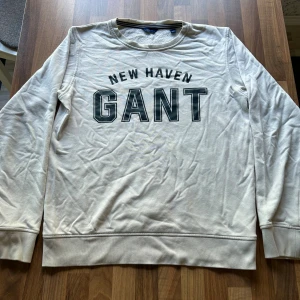 Vit sweatshirt från GANT med tryck - Säljer en vit sweatshirt från GANT med stort svart tryck 'NEW HAVEN GANT' på bröstet. Tröjan har rund hals, långa ärmar och mjuk insida. Perfekt för dig som gillar klassisk och sportig stil.