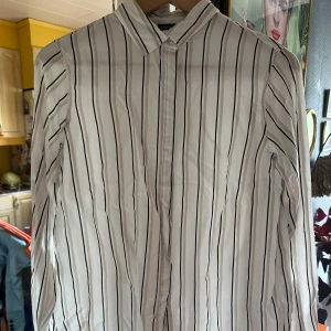 Randig skjorta från Stockh LM med silke  - Vit skjorta från Stockh LM med svarta och beige ränder. Skjortan är tunn och lätt. 34% silke & 66% viskos. Liten i strl.