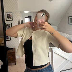 Beige bolerotopp med knapp - Säljer en stilren beige bolerotopp med korta ärmar och en snygg guldig knapp framtill. Toppen är croppad och har en öppen front, perfekt att slänga över ett linne eller topp. Materialet känns mjukt och har en lätt struktur.