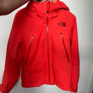 Orange/korallfärgad skidjacka från The North Face - Orange/korallfärgad skidjacka från The North Face. Bra skick, lätt vadderad, utrustad med snöbälte, skidkortsfickor, duk för att torka av skidglasögon med, innerfickor och dragkedjor med öppning för luftning vid varmare dagar. Stor luva så man får plats med hjälm under.