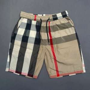 Rutigta badshorts med snörning - Snygga badshorts med klassiskt rutigt mönster i beige, svart, vitt och rött. De har elastisk midja med svart snörning och en bakficka med röd detalj. Perfekta för sommaren och har en modern, normal passform.