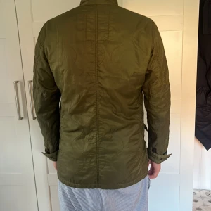 Olivgrön quiltad jacka från Ralph Lauren - Snygg olivgrön quiltad jacka från Ralph Lauren med coola detaljer som patch med amerikanska flaggan på ärmen och R.Lauren-tygmärke på bröstet. Jackan har fyra fickor med knappar, hög krage och justerbara ärmslut. Perfekt för dig som gillar utility-vibe och streetstyle.