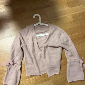 Ljusrosa omlottkofta från Lindex - Superfin ljusrosa omlottkofta från Lindex med långa ärmar och söta rosetter vid ärmsluten. Koftan är stickad och har en mjuk känsla, perfekt för dig som gillar gulliga detaljer och en chill look. Passar som en XS