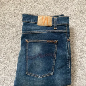 Nudie Jeans blå jeans W34 L30 - Snygga blå jeans från Nudie Jeans med klassisk femficksdesign och orangea kontrastsömmar. Jeansen har en mörk tvätt med lätt slitning och Nudie-logga på bakfickan samt läderpatch i midjan. Tillverkade i bomull och har en rak passform.
