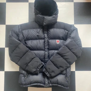 Svart dunjacka från Fjällräven S - Svart dunjacka från Fjällräven med klassisk röd logga på bröstet. Jackan har huva med snörning, två dragkedjefickor och breda quiltade paneler. Perfekt för kalla dagar och riktigt skön passform. Materialet är slitstarkt och jackan är vadderad för extra värme. Liten skada på vänster armen där är ett hål men en lapp för, går enkelt att sy ihop. Nypris är 5000 men de går ej att köpa nya längre. Det är en kamas expedition II