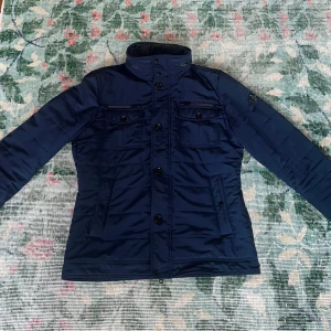 J Lindeberg fieldjacket - Snygg lite tjockare fieldjacket från J.Lindeberg som är perfekt nu under hösten och som med en tjocktröja under klarar sig en bra bit in på vintern också! Jackan har slutat produceras och finns nu endast begagnad eller liknande, men såldes ny för runt 4000. Jackan har en luva som man kan dra ut om det börjar regna via en dragkedja i nacken. Allmänt riktigt snygg och stilren jacka som passar till allt. Storlek L. Mörkblå. Supersnygg. Inga tecken på användning. Skriv vid vilken fråga som helst! ☀️