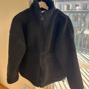 Svart teddyjacka Carin Wester XS - Mysig svart teddyjacka från Carin Wester i storlek XS. Jackan har hög krage, hel dragkedja framtill och är tillverkad i mjuk polyester med fodrad insida. Perfekt för kyliga dagar och har en loose passform med långa ärmar.