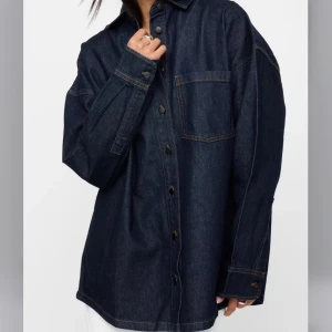 Oversized jeansskjorta i mörk denim - Säljer en oversized jeansskjorta i mörkblå denim med klassisk krage, bröstficka och knappar framtill. Skjortan har långa ärmar med manschett och kontrastsömmar som ger en snygg detalj. Aldrig använd med prislapp kvar! 