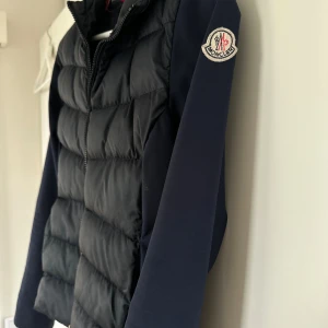 Pufferjacka från Moncler - Snygg mörk blå pufferjacka från Moncler med quiltad front och släta ärmar. Jackan har dragkedja framtill, Moncler-logga på ärmen och mjukt foder på insidan. Perfekt för dig som vill ha en stilren och varm jacka med exklusiv känsla. Lappen inuti har dessvärre gått upp i stygnen men går att fixa om man vill. Därav billigt pris. 