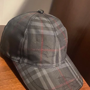 Rutig keps från Burberry i skinn - Snygg keps från Burberry i klassiskt rutigt mönster med svarta, gråa och röda toner. Yttertyget är i äkta skinn och fodret i bomull. Kepsen har broderad Burberry-logga och är tillverkad i Italien. Perfekt accessoar för en trendig streetstyle.