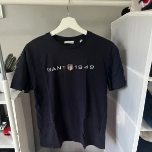 Mörkblå GANT t-shirt med tryck - Säljer en mörkblå t-shirt från GANT i storlek S. T-shirten har klassisk passform, rund hals och ett stilrent tryck med GANT 1949 och sköld på bröstet. Tillverkad i mjuk bomull som känns skön mot huden. Perfekt för en clean och enkel look. Inga defekter