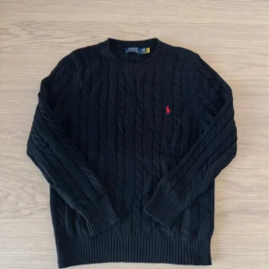 Svart kabelstickad tröja Polo Ralph Lauren - Svart kabelstickad tröja från Polo Ralph Lauren med klassisk röd logobrodyr på bröstet. Rund hals och ribbade muddar vid ärmslut och nederkant. Perfekt för dig som gillar stilren och tidlös design med en sportig touch.