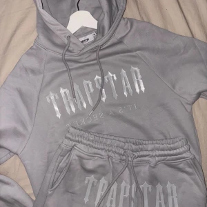 Trapstar dress - Säljer en grå hoodie från Trapstar med stort vitt tryck på bröstet och snörning i huvan. Tröjan har en klassisk passform och är tillverkad i mjukt material, perfekt för chill dagar. Snygg streetwear-vibe och passar till allt.