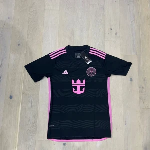 Svart och rosa Inter Miami fotbollströja - Snygg Inter Miami fotbollströja från Adidas i svart med rosa detaljer och ränder på axlarna. Tröjan har klubbmärke på bröstet, rosa logotyp och mönstrade vågor på framsidan. Tillverkad i lätt och ventilerande polyester, perfekt för match eller träning.