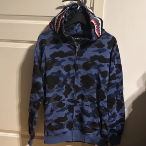 Bape hoodie - Säljer en ikonisk blå camo shark hoodie från BAPE står storlek XXL men passar M-L. Hoodien har hel dragkedja, broderade detaljer och den klassiska hajdesignen på huvan. Färgerna är blå, svart och inslag av rött och vitt. Perfekt statement-plagg för streetwear.
