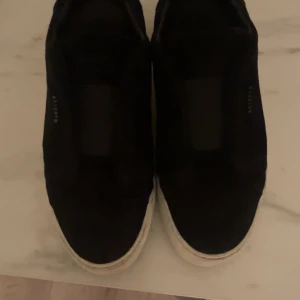 Svarta sneakers från ARIGATO - Svarta sneakers från ARIGATO med vit sula. Skorna har en stilren och enkel design utan snörning, perfekt för en clean look. Ovandelen ser ut att vara i mocka och sulan är platt och vit, vilket ger en snygg kontrast.