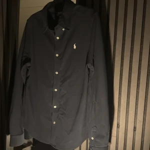Mörkblå skjorta från Polo Ralph Lauren - Snygg mörkblå skjorta från Polo Ralph Lauren med klassisk krage och vita knappar. Ikoniska Polo-loggan broderad på bröstet. Skjortan har lång ärm och är tillverkad i mjuk bomull som känns skön mot huden. Perfekt för dig som gillar stilrena och tidlösa plagg. Använd fåtal gånger!