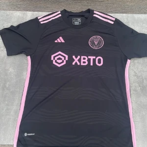 Inter Miami svart fotbollströja Adidas L - Svart Inter Miami CF fotbollströja från Adidas i storlek L, slim fit. Tröjan har rosa detaljer som logga, ränder på axlarna och sponsortryck. Materialet är lätt och andas, perfekt för träning eller match. Rund hals och korta ärmar, med subtila vågmönster i tyget.