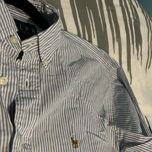 Randig skjorta från Ralph Lauren - Snygg skjorta från Ralph Lauren med blå och vita ränder, klassisk krage och knappar framtill. Broderad logga på bröstet i guld. Tillverkad i 100% bomull, perfekt för dig som gillar stilrena och tidlösa plagg. Långärmad modell med relaxed fit.