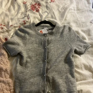 Grå kortärmad kofta från H&M - Mysig grå kofta från H&M med korta ärmar och rund hals. Koftan är stickad i mjukt material och har knappar framtill. Perfekt att slänga över en topp för en chill look. Passar dig som gillar enkel och stilren design.