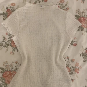 Vit strukturerad topp från Kappahl - Säljer en söt vit topp från Kappahl i storlek XS. Toppen har korta ärmar, lätt rynkad struktur och volangkant vid hals och ärmslut. Perfekt att styla med jeans eller kjol för en fräsch look.
