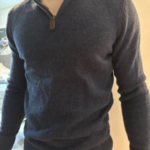 Mörkblå stickad tröja med half zip - Mörkblå stickad tröja med half zip och ribbade muddar. Tröjan har lång ärm och en klassisk passform som sitter snyggt på kroppen. Perfekt för kyligare dagar och enkel att matcha med jeans eller chinos.