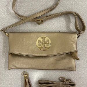 Ny lyxig axelväska från Tory Burch - Ny snygg champagnefärgad axelväska från Tory Burch med justerbar rem och stort logomärke i metall framtill. Tillhörande tillbehör: extra axelband och tofs. Väskan har dragkedja, extra fack på baksidan och insida med mönstrat tyg i brunt och beige. Perfekt storlek för det viktigaste och stilren design. Fått i present, kan ej garantera original. Skickas eller hämtas i centrala Göteborg.