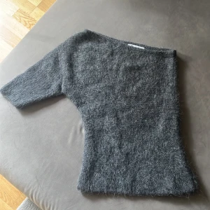 Grå stickad oneshoulder topp från Zara - Säljer en grå stickad oneshoulder topp från Zara i storlek M. Toppen har en asymmetrisk design med en ärm och är tillverkad i ett fluffigt stickat material som känns mjukt och cozy. Perfekt för dig som gillar unika silhuetter och vill sticka ut lite extra.