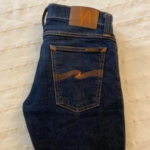 Mörkblå jeans från Nudie Jeans Co - Snygga mörkblå jeans från Nudie Jeans Co med klassisk orange söm och läderpatch bak i midjan. Jeansen har raka ben och är tillverkade i slitstarkt bomullsjeans. Perfekt passform och stilren look med tydliga Nudie-detaljer.
