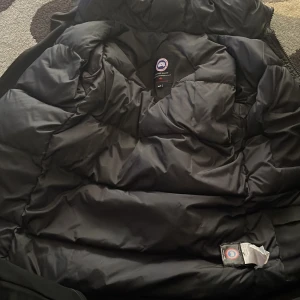 Svart dunjacka Canada Goose med pälskrage - Säljer en svart dunjacka från Canada Goose med klassisk patch på ärmen och huva med fluffig pälskant. Jackan är lång, har dragkedja och knappar framtill samt flera praktiska fickor. Perfekt för kalla vinterdagar och riktigt snygg streetstil.