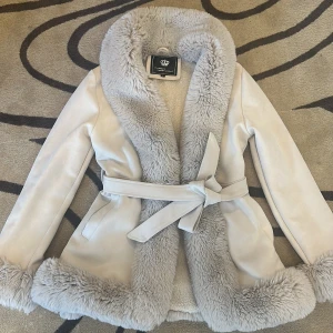 Beige pälsjacka med bälte S -  en supermjuk och fluffig beige jacka med bred krage och muddar i fuskpäls. Den har bälte i midjan för en snygg passform och ett mjukt, mysigt material perfekt för kalla dagar när man vill hålla sig både varm och snygg.