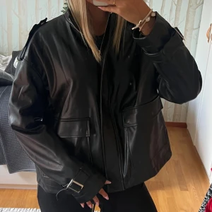 Svart oversized skinnjacka med detaljer - Säljer en svart oversized skinnjacka med coola spänndetaljer vid ärmsluten och stora fickor framtill. Jackan har dragkedja och en rak, boxig passform som ger en modern vibe. Perfekt för dig som vill ha en edgy look till din outfit. Från nakd passar xs-m beroende på hur du vill att den ska sitta❤️