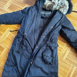 Svart parkasjacka med fuskpäls från The Smirk - Svart parkasjacka från The Smirk med huva och fuskpäls i gråbruna toner. Jackan har dragsko i midjan, stora fickor med lock och dragkedja framtill. Fodrad insida i quiltad polyester gör den perfekt för kalla dagar.