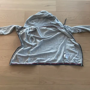 Grå hoodie från Tommy Hilfiger - Säljer en grå hoodie från Tommy Hilfiger med huva och dragkedja. Tröjan har snyggt band med logga längs nederkanten. Tillverkad i mjuk bomullsmix och har en liten broderad logga på bröstet. Perfekt för chill dagar.