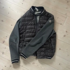Grå Moncler cardigan - Säljer en snygg cardigan från Moncler i storlek S. Jackan har quiltad svart front med grå ärmar och rygg, samt randiga muddar vid ärmslut och midja. Dragkedja framtill och klassisk Moncler-logga på ärmen. Perfekt för höst och vår. Passar runt 170cm.