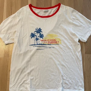 Vit Saint Laurent t-shirt med tryck - Vit t-shirt från Saint Laurent med röd krage och tryck i blått och gult framtill med texten 'Waiting for Sunset' och palmer. Skön och avslappnad passform, perfekt för sommardagar. Tillverkad i mjuk bomull. Fläcken går att ta bort