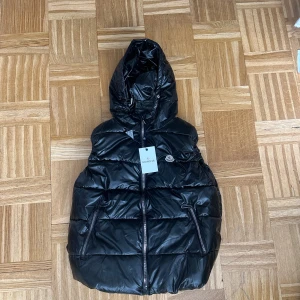 Svart pufferjacka från Moncler - Snygg svart pufferjacka från Moncler med glansig finish och huva. Jackan har två dragkedjeförsedda fickor och en Moncler-logga på bröstet. Perfekt för kalla dagar och har en modern, oversized look.