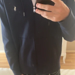 Mörkblå hoodie från Ralph Lauren - Säljer en mörkblå hoodie från Ralph Lauren med dragkedja och huva. Tröjan har den klassiska vita Polo-loggan på bröstet, snörning i huvan och stora fickor framtill. Perfekt för dig som gillar stilren och sportig look.