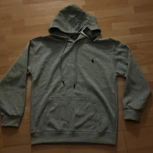 Grå hoodie från Polo Ralph Lauren - Säljer en rikigt snygg hoodie från polo Ralph lauren. Den är helt oanvänd med prislapp och påse som med följer. Skulle du ha några andra frågor eller funderingar är de bara att höra av sig!