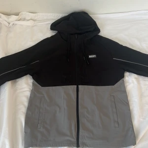 Svart och grå vindjacka från McKenzie - Säljer en svart och grå vindjacka från McKenzie i storlek M. Lite smutsig, men det kan komma av med lite vatten eller i tvätten. Jackan har huva med snörning, hel dragkedja framtill och två sidofickor. Perfekt för blåsiga dagar och enkel att matcha med streetwear. Materialet är lätt och känns smidigt.