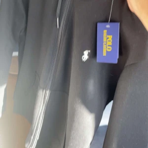 Svart hoodie från Polo Ralph Lauren - Svart hoodie från Polo Ralph Lauren med dragkedja framtill och klassisk vit logga på bröstet. Tröjan har huva och fickor på sidorna, perfekt för en chill och stilren look.