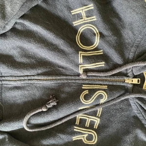 Mörkgrå hoodie från Hollister - Mörkgrå hoodie från Hollister med dragkedja och snörning i huvan. Stor logga i vitt framtill. Skön och avslappnad passform, perfekt för chill dagar. Klassisk streetstyle-vibe och mjukt material.
