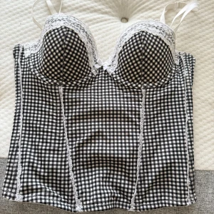 Rutig korsettopp från H&M med spets - Säljer en svartvit rutig korsettopp från H&M i storlek 38. Toppen har vadderade kupor, vita spetsdetaljer och en liten rosett framtill. Axelbanden är vita och tyget är mjukt med stretch. Perfekt för att lyfta din outfit med en söt och trendig touch. Hör av dig vid funderingar💕